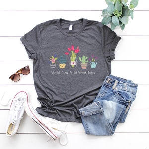 May include: A dark heather grey t-shirt with a graphic of potted plants and the text "We All Grow At Different Rates".