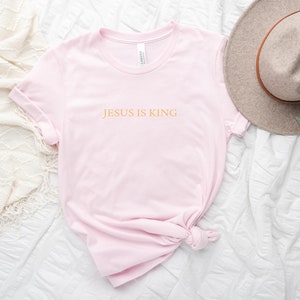Pu&ograve; includere: Una maglietta rosa con il testo "JESUS IS KING" in lettere dorate.
