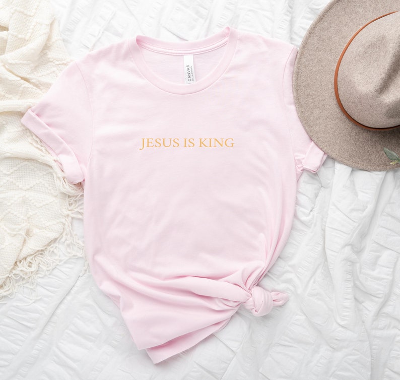 Pu&ograve; includere: Una maglietta rosa con il testo "JESUS IS KING" in lettere dorate.