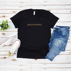 Pu&ograve; includere: Maglietta nera con il testo "JESUS IS KING" in lettere dorate. La maglietta &egrave; fotografata su uno sfondo di legno bianco con un paio di scarpe da ginnastica bianche, jeans blu e una piccola pianta verde.