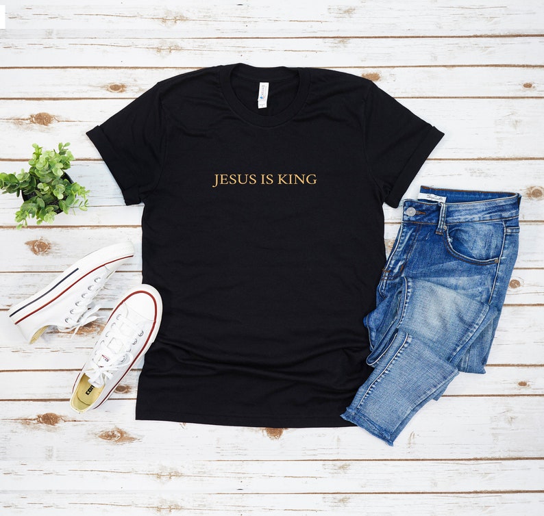 Pu&ograve; includere: Maglietta nera con il testo "JESUS IS KING" in lettere dorate. La maglietta &egrave; fotografata su uno sfondo di legno bianco con un paio di scarpe da ginnastica bianche, jeans blu e una piccola pianta verde.