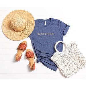 Pu&ograve; includere: Una maglietta blu navy con il testo "JESUS IS KING" in lettere dorate. La maglietta &egrave; stesa su una superficie bianca con un cappello di paglia, sandali e una borsa di crochet bianca.