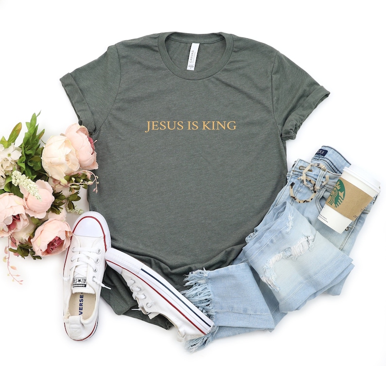Pu&ograve; includere: Una maglietta verde scuro con il testo "JESUS IS KING" in lettere dorate. La maglietta &egrave; su uno sfondo bianco con un paio di scarpe da ginnastica bianche, jeans blu e una tazza di caff&egrave;.
