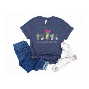 May include: A navy blue t-shirt with a graphic of potted plants and the text "We All Grow At Different Rates". The shirt is paired with blue jeans and white sneakers.