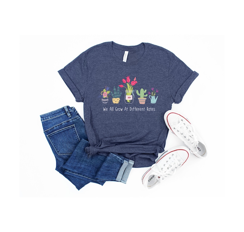 May include: A navy blue t-shirt with a graphic of potted plants and the text "We All Grow At Different Rates". The shirt is paired with blue jeans and white sneakers.