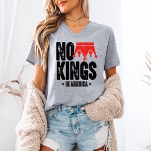 A woman wearing a grey t-shirt with the text "NO KINGS IN AMERICA" printed on it, standing in a room with a beige sweater draped over her shoulders.