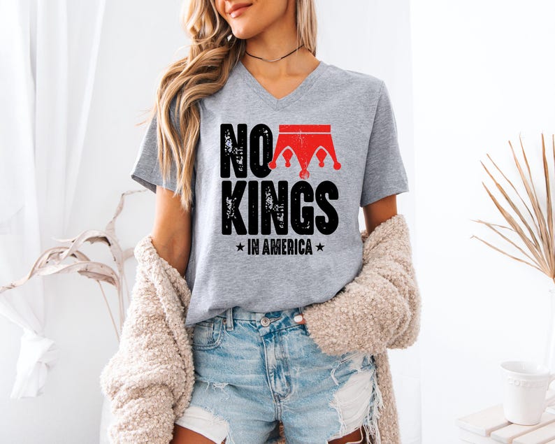 A woman wearing a grey t-shirt with the text "NO KINGS IN AMERICA" printed on it, standing in a room with a beige sweater draped over her shoulders.