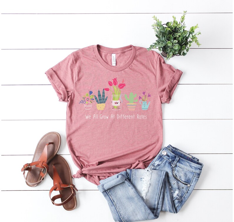 May include: A pink t-shirt with a graphic of potted plants and the text "We All Grow At Different Rates".