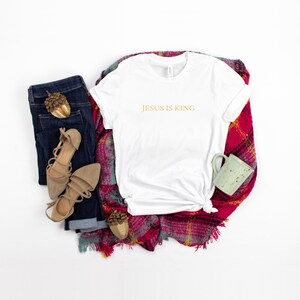 Pu&ograve; includere: Maglietta bianca con il testo "JESUS IS KING" in lettere dorate. La maglietta &egrave; appoggiata su una coperta a quadri rossi e bianchi con un paio di sandali marroni, un paio di jeans blu e una tazza verde.