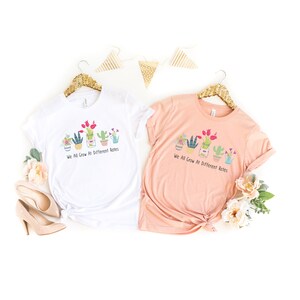 May include: Two t-shirts with a graphic of potted plants and the text "We All Grow At Different Rates". One shirt is white and the other is peach.
