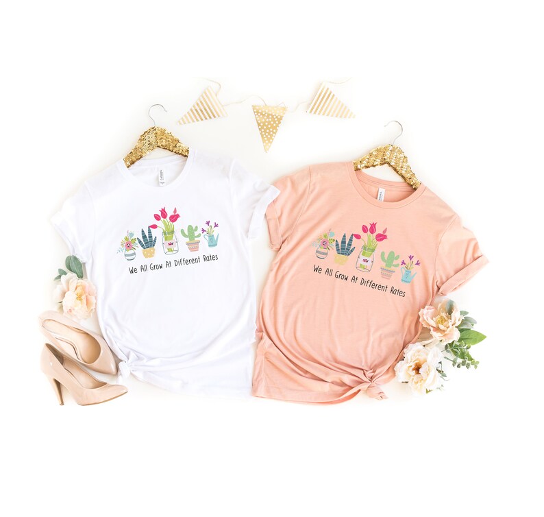 May include: Two t-shirts with a graphic of potted plants and the text "We All Grow At Different Rates". One shirt is white and the other is peach.