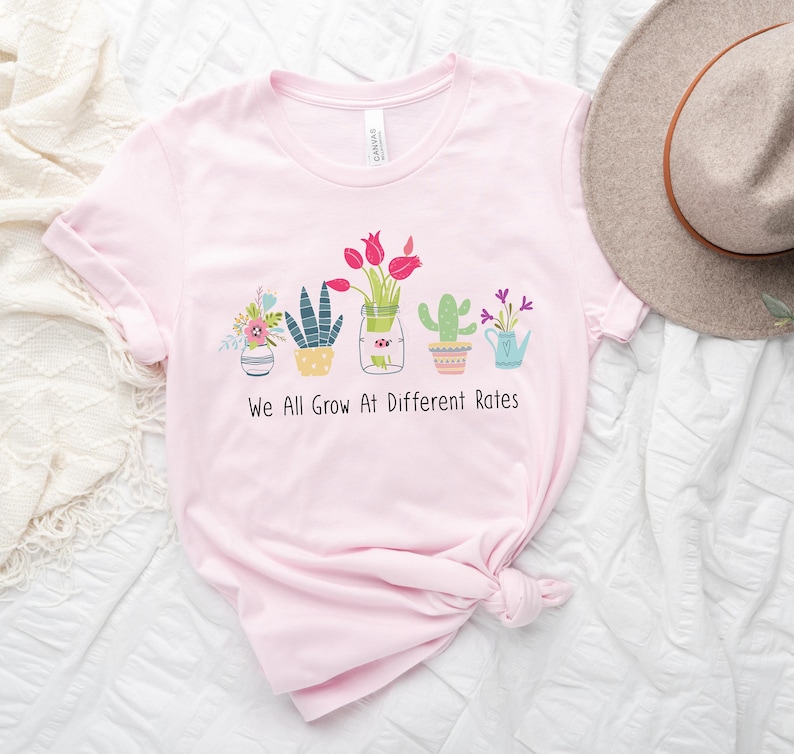 May include: Pink t-shirt with a graphic of potted plants and the text "We All Grow At Different Rates".