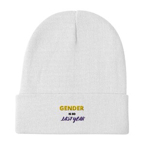 Op de afbeelding: Witte gebreide beanie met de tekst "GENDER IS SO LAST YEAR" geborduurd in geel en paars.