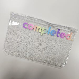 May include: Transparent rectangular pencil case with a silver glitter effect. The word "completed!" is written across the top in rainbow-coloured letters. The case has a zip closure and small holes along the bottom edge.