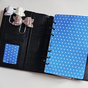 May include: Black planner with a blue polka dot insert and matching card. Three decorative bow paper clips in white, light pink glitter, and pale pink glitter are attached to the planner.