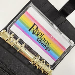 May include: Black faux leather binder with gold-coloured metal rings. A clear plastic insert displays a rainbow-striped design with the text "RBPlans savings". The binder is open, revealing the interior.
