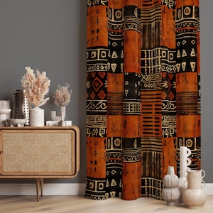 Könnte beinhalten: Gemusterter Vorhang mit Patchwork-Design. Der Vorhang zeigt geometrische Formen und Stammesmuster in Schwarz, Beige und Orange. Der Vorhang hängt vor einer grauen Wand. Links befindet sich ein Holzschrank mit Vasen und getrockneten Pflanzen.