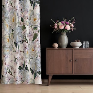 Könnte beinhalten: Ein bodenlanger Vorhang mit einem sich wiederholenden Muster aus großen weißen und hellblauen Rosen mit grünen Blättern und kleinen rosa Blüten auf cremefarbenem Grund, der neben einem dunkelbraunen Holzschrank mit einer Blumenvase hängt.