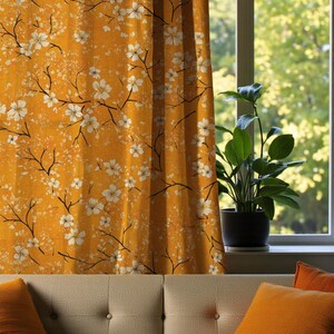 Könnte beinhalten: Senfgelber Vorhang mit einem floralen Muster aus weißen Blüten und schwarzen Zweigen. Der Vorhang hängt vor einem Fenster mit Blick ins Grüne. Im Vordergrund befindet sich ein beiges Sofa mit orangefarbenen Kissen.