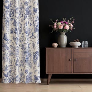 Könnte beinhalten: Ein bodenlanger Vorhang mit blau-weißem botanischem Blumenmuster neben einem dunkelbraunen Holzschrank. Auf dem Schrank stehen eine graue Keramikvase mit rosa und weißen Blumen, eine kleine Schale und ein klares Glas.