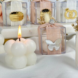 May include: Clear plastic boxes with white and pink ribbon, filled with small white candies. The boxes are decorated with dried flowers, gold accents, and personalized name tags. A white candle with a bubble design is in the foreground.