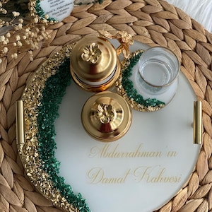 May include: A white ceramic tray with gold and green accents. The tray is decorated with gold leaf and green stones. The tray has a handle on the side and is inscribed with the text "Abdurrahman'in Damat Kahvesi".