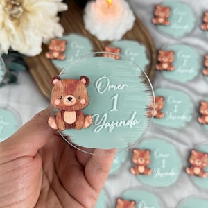 May include: A clear, round acrylic favor with a light teal background and a cartoon bear. The text "Ömer 1 Yaşında" is written in white script. Several similar favors are visible in the background.