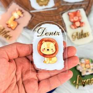 May include: A clear acrylic name tag with a white background and a gold inscription that reads "Deniz". A cartoon lion with a brown mane and a yellow body is sitting on the tag.