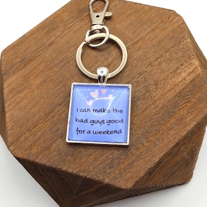 Può includere: Un portachiavi quadrato con una cornice in metallo color argento e uno sfondo blu. Il portachiavi ha il testo "I can make the bad guys good for a weekend" in nero. Due cuori rosa sono sopra il testo. Il portachiavi è attaccato a un anello e un fermaglio in metallo color argento.
