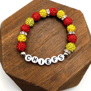 Könnte beinhalten: Ein Armband mit roten und gelben glitzernden Perlen, durchsetzt mit silbernen Abstandshaltern. Das Armbandbuchstabiert "CHIEFS" in weißen Buchstabenperlen. Das Armband wird auf einer Holzoberfläche präsentiert.
