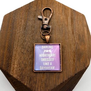 Può includere: Un portachiavi quadrato con cornice e clip color rame. Il centro presenta uno sfondo viola e blu con il testo "Darling I'm a Nightmare Dressed Like a Daydream" in bianco. Il portachiavi è esposto su una superficie di legno.
