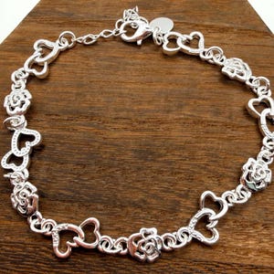 May include: Silver-toned bracelet featuring alternating heart and rose-shaped charms. The bracelet is made of linked metal and has a lobster clasp closure. The bracelet is displayed on a wooden surface.