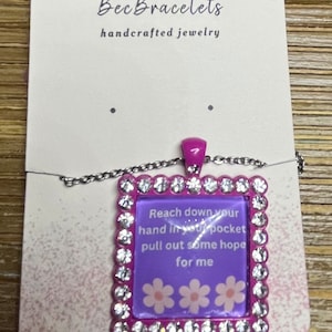 May include: A pink square pendant necklace with rhinestones around the edge. The pendant has a purple background with white flowers and the text "Reach down your hand in your pocket pull out some hope for me".