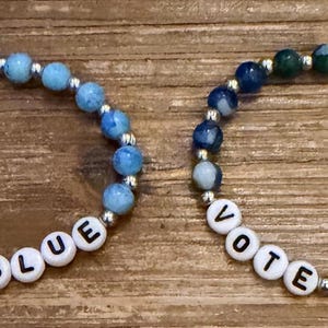 May include: Two beaded bracelets. One features light blue and silver beads, the other has dark blue and green beads. Both bracelets spell out "VOTE BLUE" with white letter beads. The bracelets are on a wooden surface.