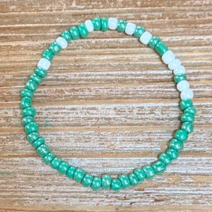 May include: A circular beaded bracelet constructed from small, round, glossy beads. The design features a repeating pattern of vibrant green beads interspersed with single white beads, creating a simple, continuous loop of colourful, uniform glass-like spheres.