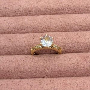 May include: A gold-colored ring with a large, round, clear gemstone as the centerpiece. The band is detailed with smaller gemstones. The ring is set against a pink, textured background.