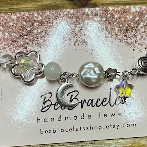 May include: A silver charm bracelet with a moon, stars, and a flower charm. The bracelet is on a pink background with the text "Bee Bracel handmade jewelry becbraceletshop.etsy.com".