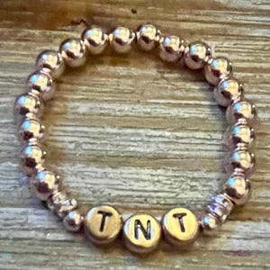 Könnte beinhalten: Ein roségoldenes Perlenarmband mit den Buchstaben "TNT" in goldenen Buchstabenperlen. Das Armband besteht aus kleinen, runden Perlen und liegt auf einer Holzoberfläche.