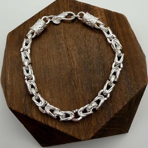 May include: A silver bracelet with a detailed chain design. The bracelet features a series of interlocking links, with decorative cylindrical end pieces. The bracelet is displayed on a dark wooden surface.