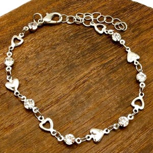 May include: Silver-toned bracelet featuring heart and open-heart shaped links, interspersed with clear, round gemstones. The bracelet has an adjustable chain for a custom fit and is displayed on a wooden surface.