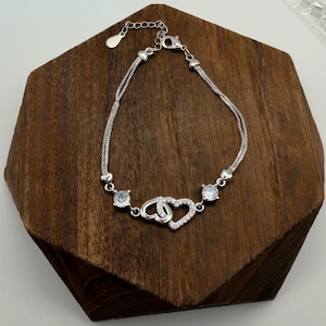 May include: A silver bracelet with two delicate chains and a heart-shaped charm. The charm features two interlocking hearts, one encrusted with small clear stones. Two clear, round stones are set on either side of the heart charm.