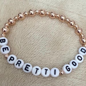 May include: A rose gold beaded bracelet with the words "BE PRETTI GOOD" spelled out in white letter beads. The bracelet is made of small, round, rose gold beads and white letter beads with black letters.
