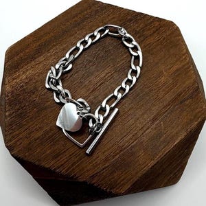 May include: A silver-toned metal chain bracelet featuring a toggle clasp closure. The bracelet includes a smooth, polished heart-shaped charm attached near the toggle bar, which is designed to slide through a circular loop to secure the piece.