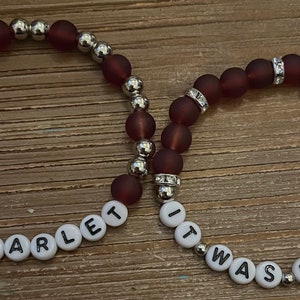 May include: Two beaded bracelets with silver beads and maroon colored beads. One bracelet spells out "So Scarlet" and the other spells out "It Was Maroon".