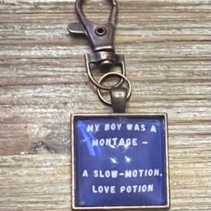 Pode incluir: Um porta-chaves de cor bronze com um pingente retangular. O pingente tem um fundo azul com o texto "MY BOY WAS A MONTAGE - A SLOW-MOTION, LOVE POTION" em branco. O porta-chaves tem um fecho de lagosta.