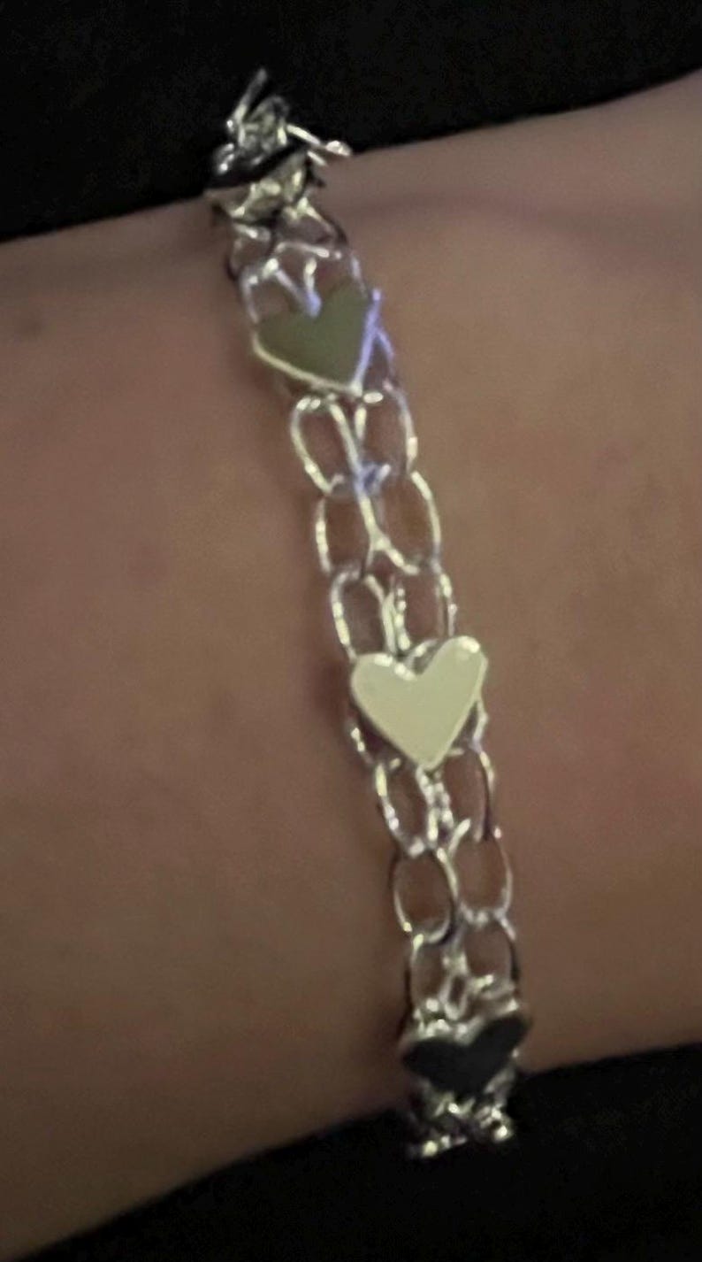 May include: A silver chain bracelet with three heart-shaped charms. The charms are silver, white, and black.