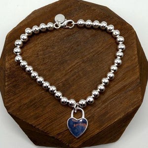 May include: A silver-toned beaded bracelet featuring a series of uniform, polished spherical beads. The bracelet includes a lobster claw clasp and a heart-shaped padlock charm with a dark, mottled blue and copper-colored surface finish.