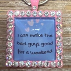 Può includere: Un portachiavi quadrato rosa con sfondo blu e la scritta "I can make the bad guys good for a weekend". Il portachiavi è incorniciato con strass trasparenti e ha un pezzo superiore rosa e un anello argentato.