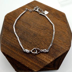 May include: A silver bracelet with a delicate design. The bracelet features a series of connected bars and two heart-shaped charms. Small, clear gemstones are set near the heart charms. The bracelet is displayed on a wooden surface.