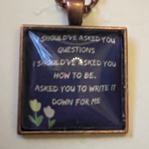 Puede incluir: Un collar con un colgante cuadrado de vidrio azul y una cadena plateada. El colgante tiene un texto blanco que dice "SHOULD'VE ASKED YOU QUESTIONS I SHOULD'VE ASKED YOU HOW TO BE. ASKED YOU TO WRITE IT DOWN FOR ME". El colgante también tiene dos flores blancas con tallos verdes.